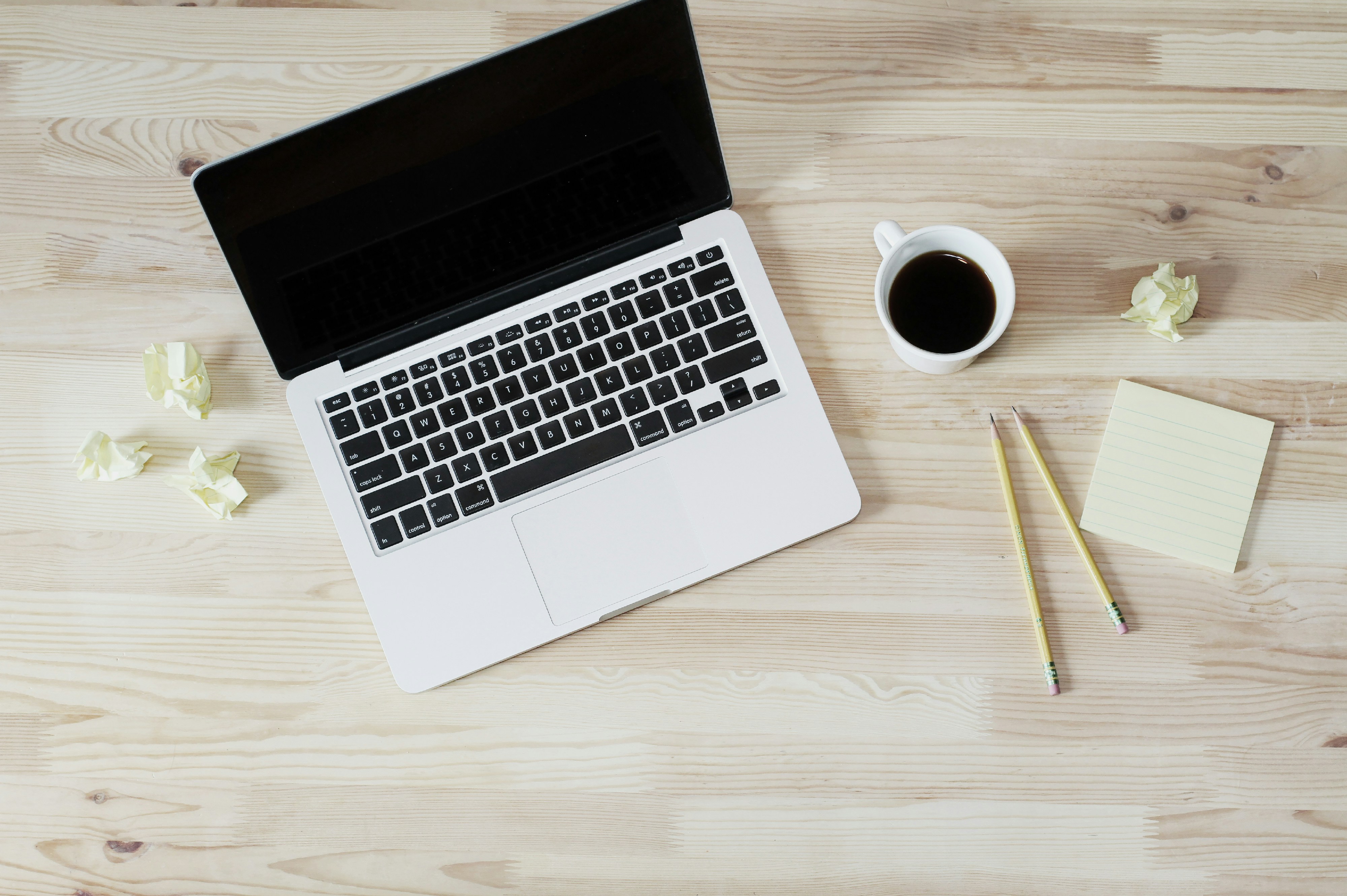 A laptop is open next to a cup of coffee and two pencils on a blond wood table.