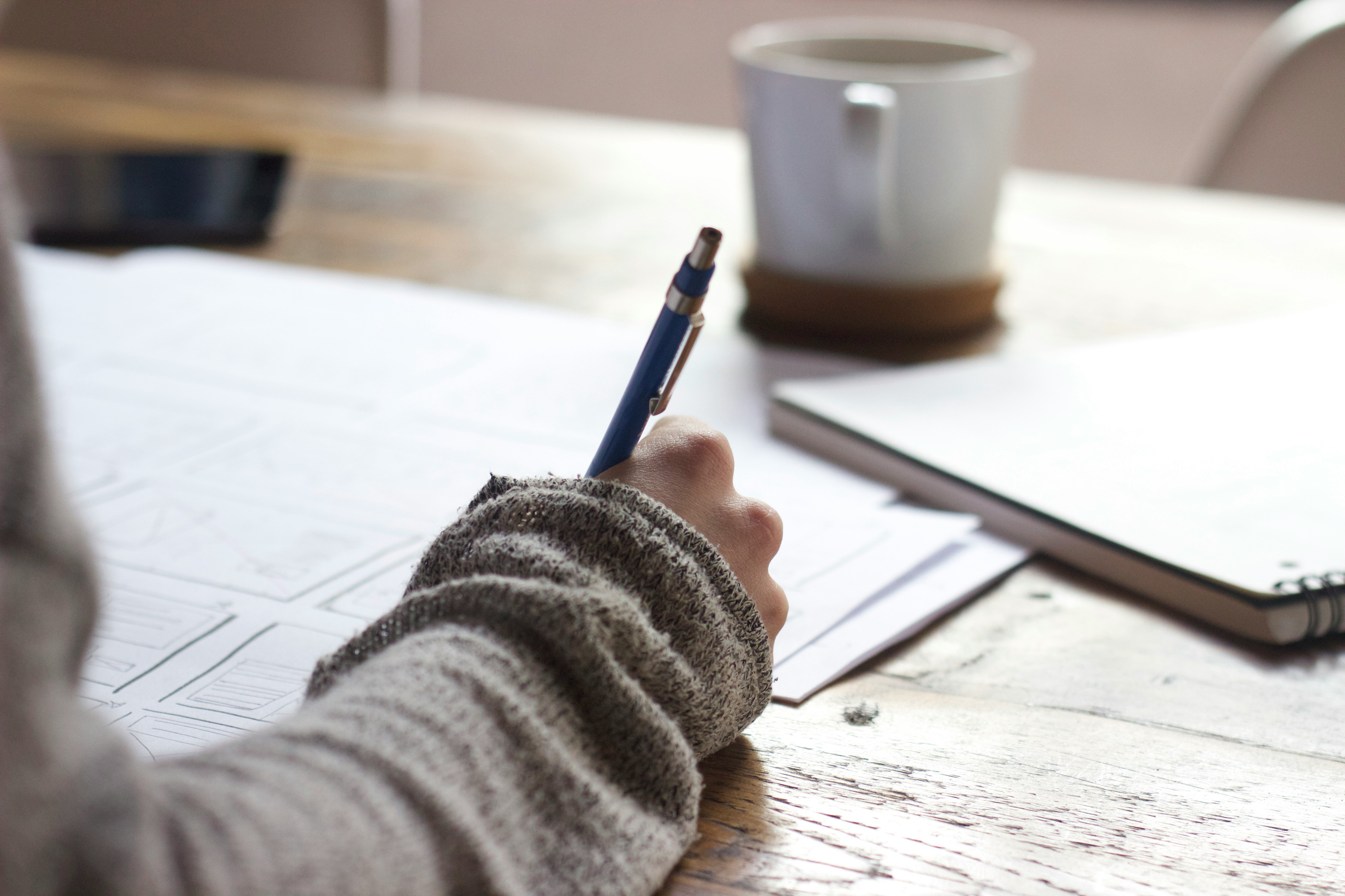 A hand holds a blue pencil and writes on paper. There is a white coffee mug in the background.